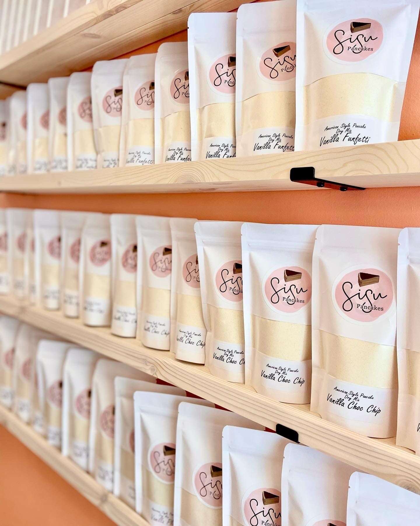 Multiple rows of packets of Sisu Pancakes dry mix arranged on light wood shelves against a peach-colored wall.