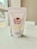 A stand-up pouch of vanilla flour with a clear window showing the pale yellow powder inside. The pouch has a white label with a pink circle and a cake slice graphic, reading 'Sisu Pancakes' and 'Vanilla Plain'.