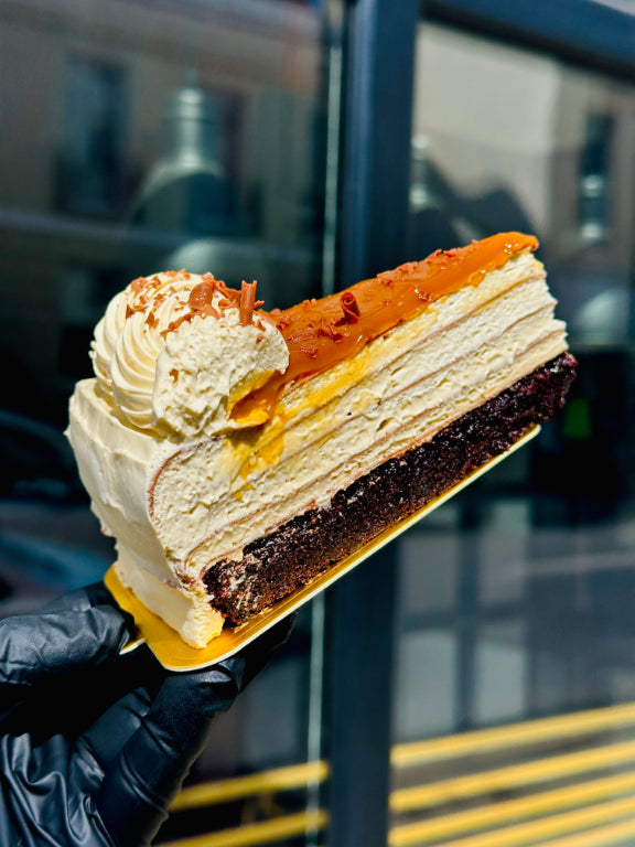 A close-up of a slice of pancake cake, held by a hand wearing a black glove. The cake has cream-colored layers, a dark chocolate base, and a glossy orange topping, with whipped cream on top.
