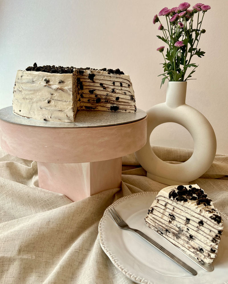 Layered cookies and cream pancake cake with a slice on a plate, next to a vase with flowers on a beige tablecloth.