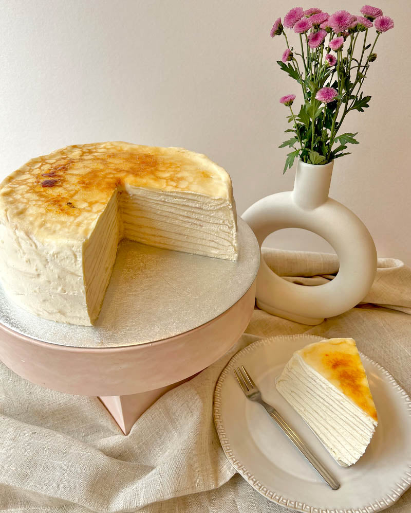 Layered creme brulee pancake cake on a stand with a slice on a plate, accompanied by a vase of flowers.