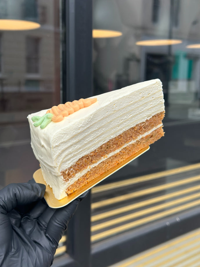 A hand wearing a black glove holds a slice of layered carrot pancake cake with white frosting on a small gold plate, with a blurred storefront window in the background.