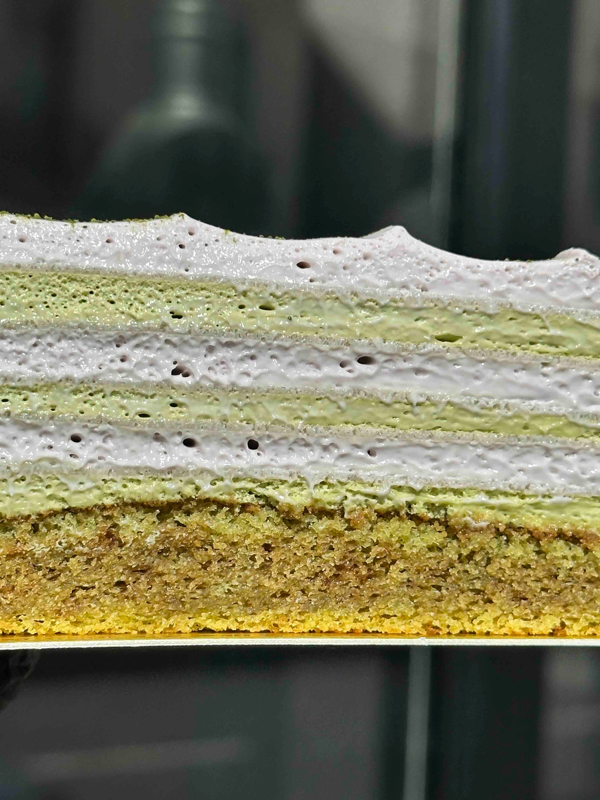 A close-up cross-section of a layered cake slice, showing alternating layers of pale green and white filling, and a golden-brown sponge base.