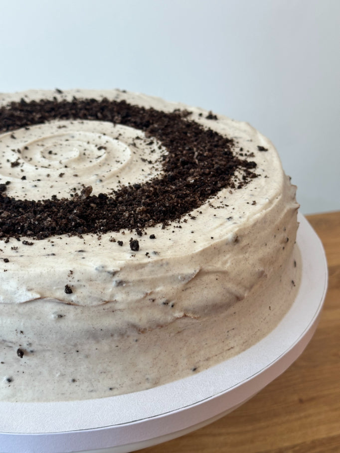 A round pancake cake with light cream frosting and a cookie crumb swirl on top, sitting on a white plate.