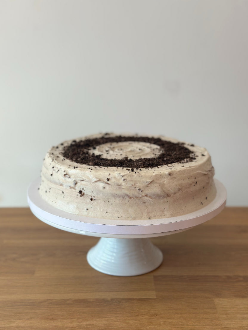 A round pancake cake with cookie crumbles on top, displayed on a white pedestal stand against a plain light wall and wooden surface.