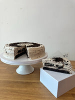 A slice of layered cookies and cream pancake cake with chocolate cookie filling and light cream frosting, displayed on a white pedestal stand on a wooden table against a plain light wall.