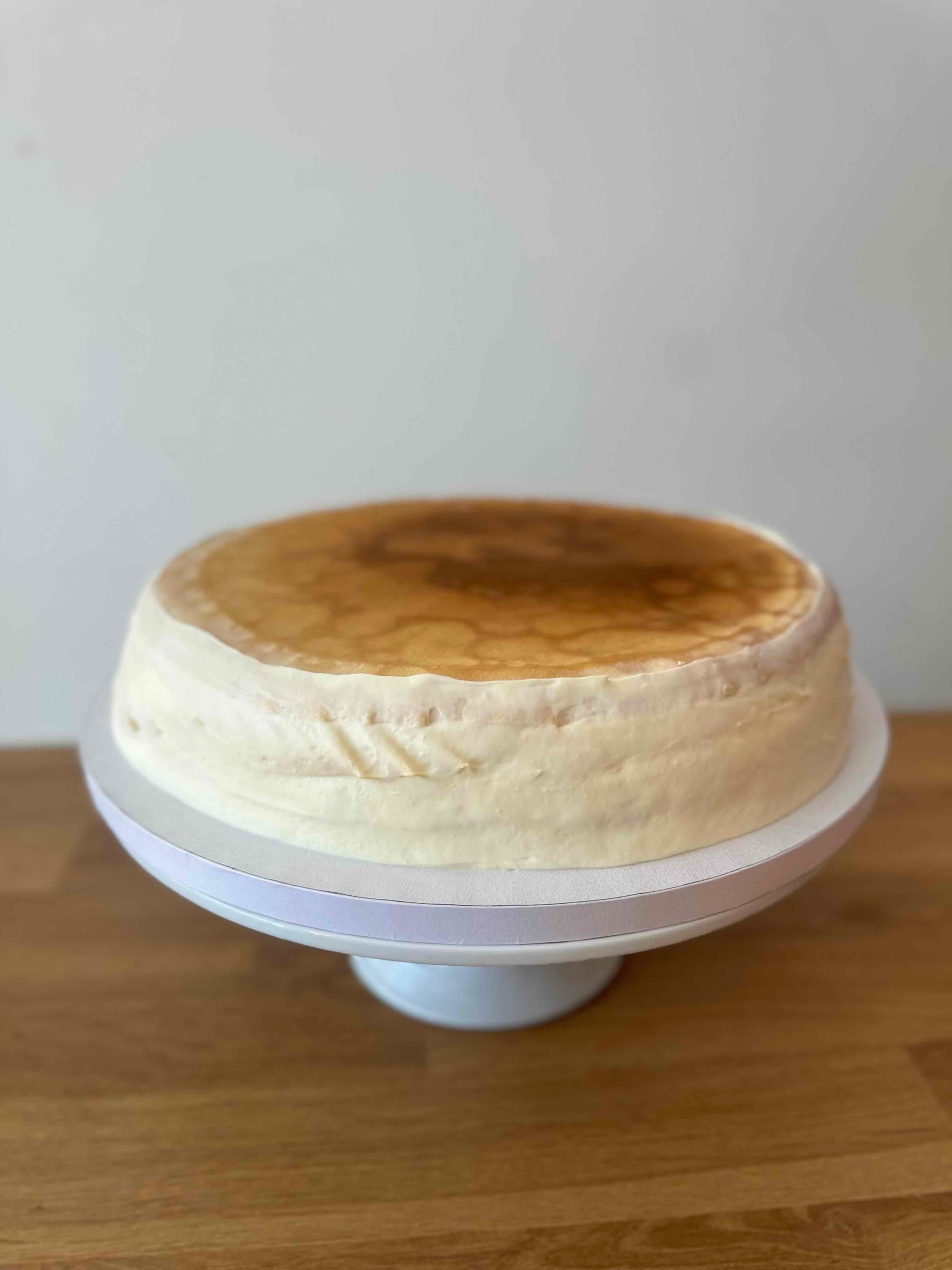 A round creme brulee pancake cake with a golden-brown top and pale cream sides, displayed on a white cake stand over a wooden surface against a plain light wall.