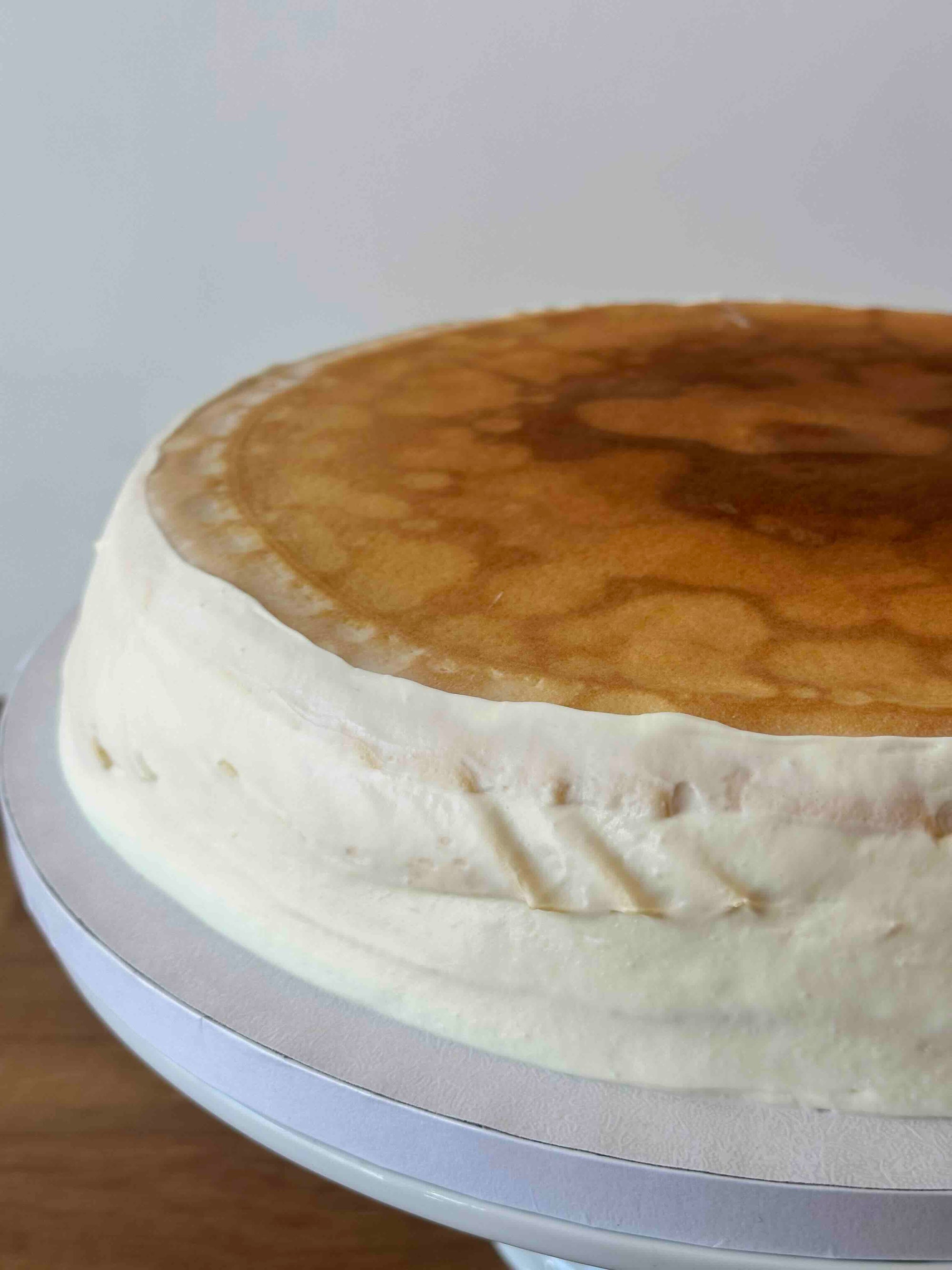A round creme brulee pancake cake with smooth off-white frosting on the sides and a golden-brown top, shown on a white cake stand against a plain light background.