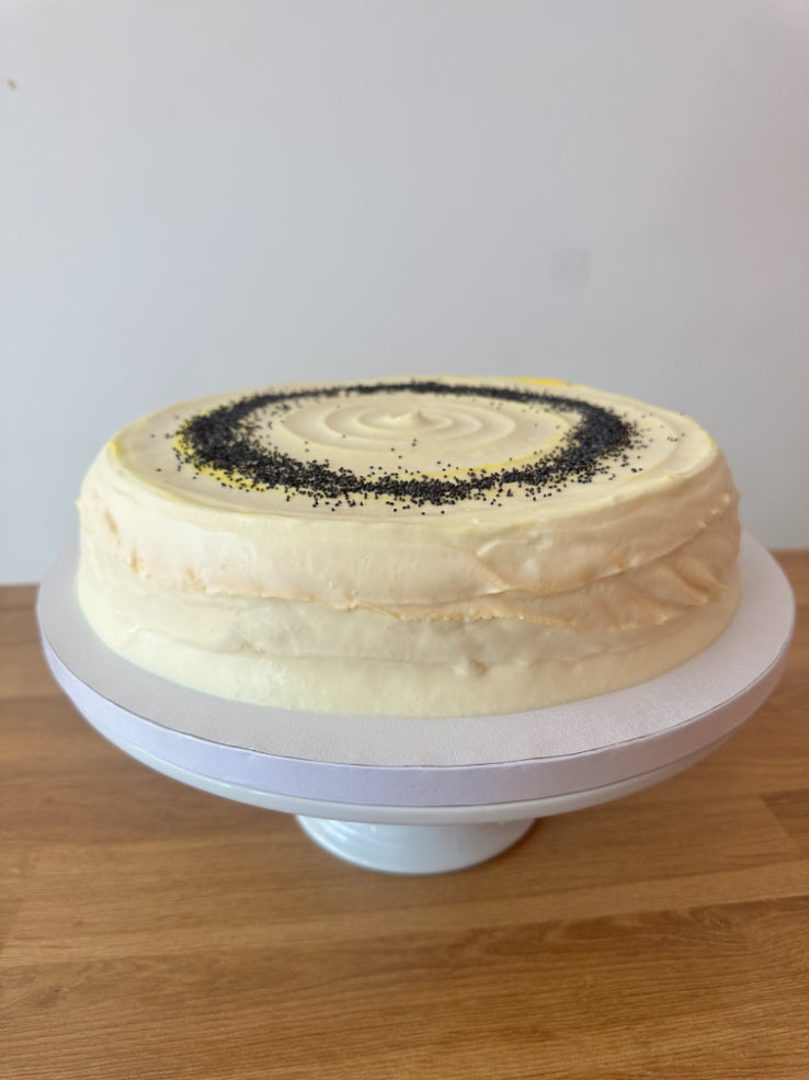 A round frosted pancake cake with pale cream icing sits on a white cake stand on a wooden table against a plain light wall. The top is decorated with a spiral pattern of poppy seeds.
