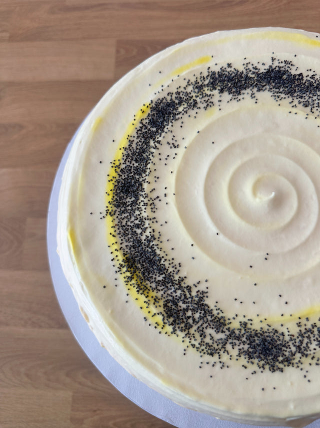 A round frosted pancake cake with white icing, decorated with a spiral pattern and a ring of black poppy seed sprinkles, shown on a wooden tabletop.