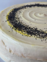 A close-up of a round pancake cake with smooth off-white frosting, decorated with a spiral pattern of dark poppy seeds and a thin ring of yellow frosting.