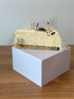 A slice of layered pancake cake with a golden-brown sponge base, displayed on a white square box on a wooden surface.
