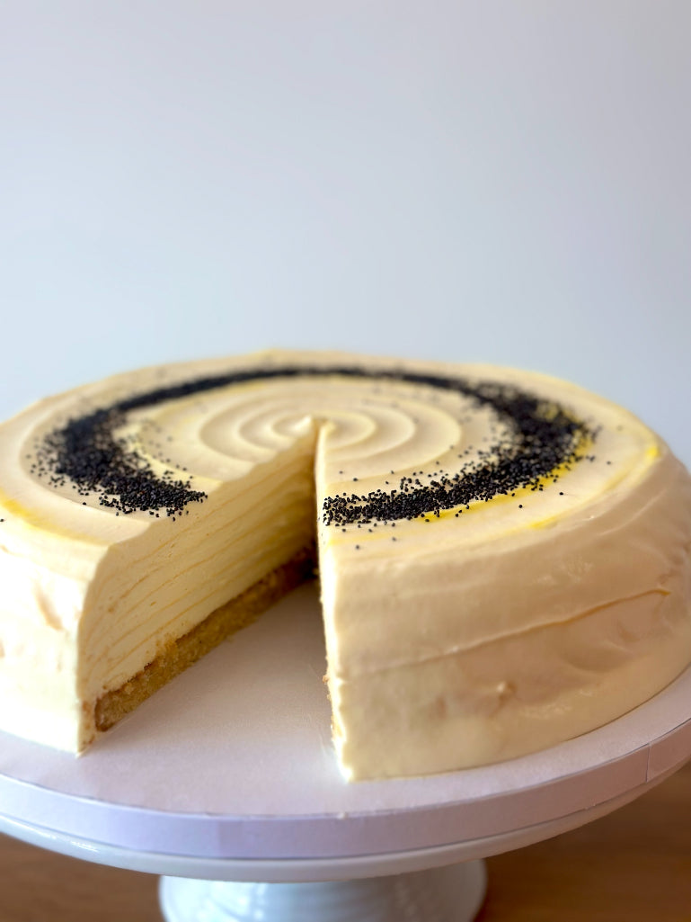 A layered pancake cake with pale cream frosting and a swirl of poppy seeds on top, shown with one slice removed on a white cake stand against a light gray background.