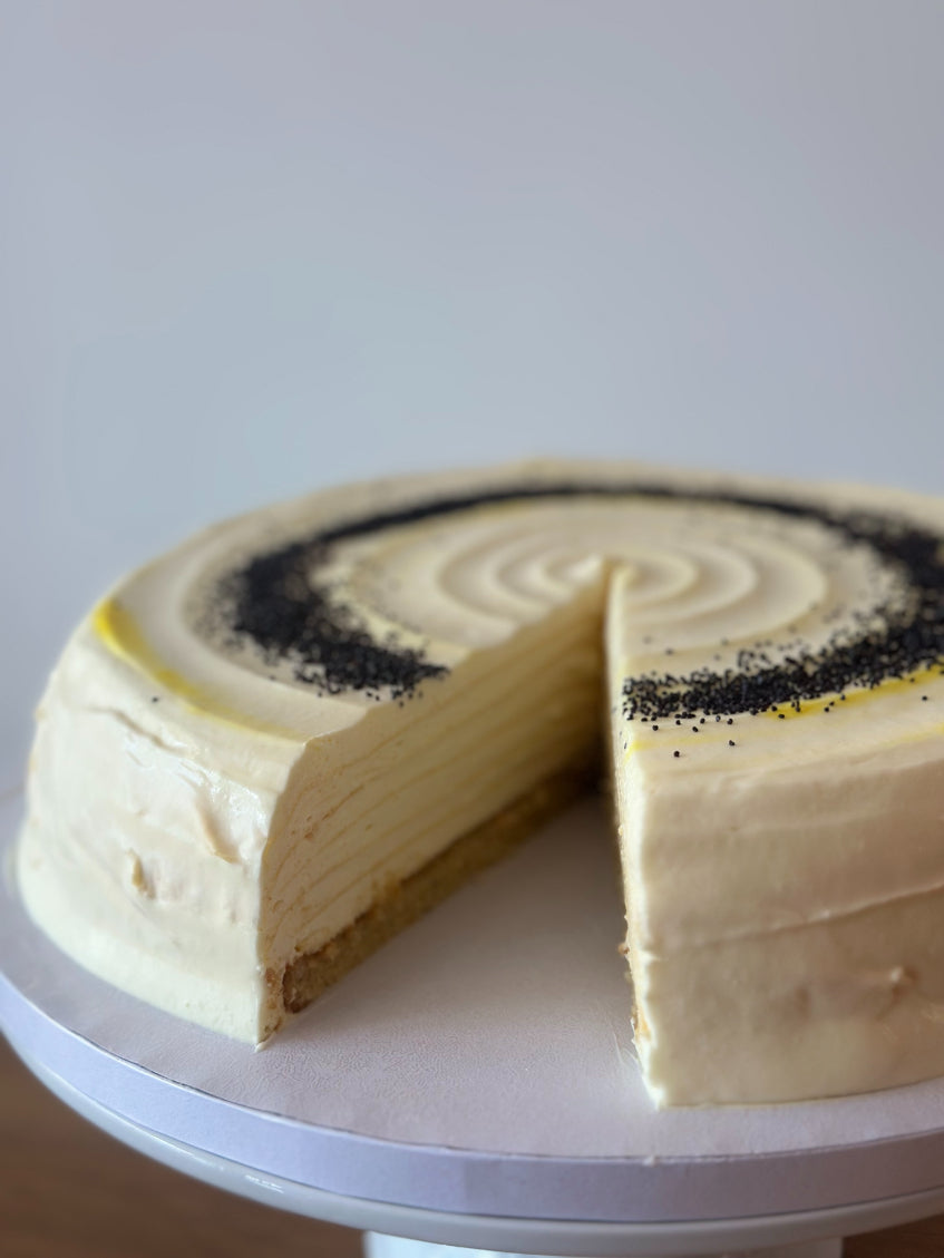 A round layered pancake cake with pale cream frosting and a dark poppy seed topping, shown on a white cake stand against a light gray background.