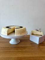 A layered pancake cake with cream-colored frosting and poppy seeds sits on a white pedestal stand, with a slice placed on a small white box beside it on a wooden table against a plain wall.