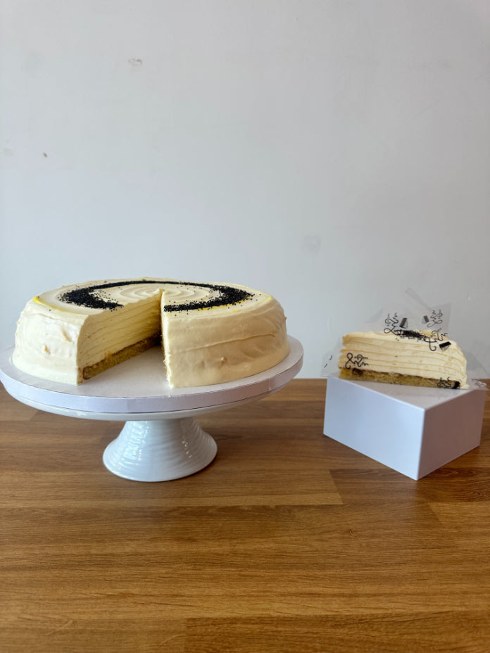 A layered pancake cake with cream-colored frosting and poppy seeds sits on a white pedestal stand, with a slice placed on a small white box beside it on a wooden table against a plain wall.