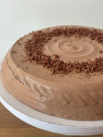 A round chocolate hazelnut pancake cake with smooth brown frosting and chocolate shavings on top, shown on a white cake board.