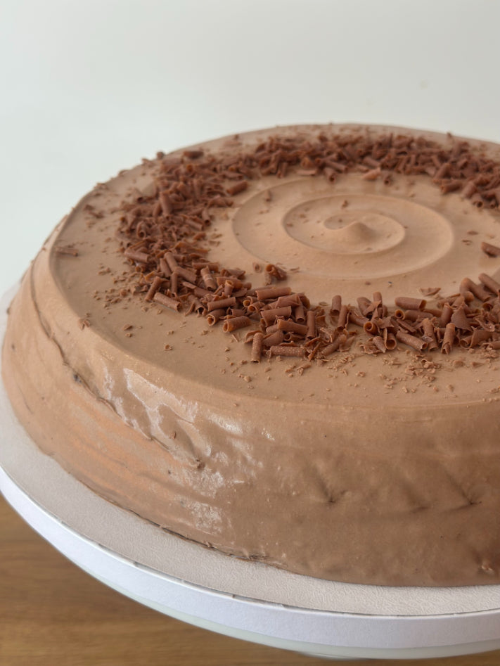 A round chocolate hazelnut pancake cake with smooth brown frosting and chocolate shavings on top, shown on a white cake board.