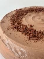 Chocolate hazelnut pancake cake with smooth chocolate frosting and chocolate shavings sprinkled on top, photographed in a close-up view against a light neutral background.