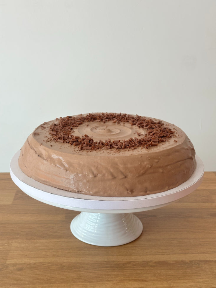 A round chocolate hazelnut pancake cake with smooth brown frosting and chocolate shavings on top, displayed on a white pedestal stand on a wooden surface against a plain light wall.