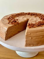 Chocolate hazelnut pancake cake with multiple layers and chocolate frosting, topped with chocolate shavings, shown on a white cake stand with a slice cut out.