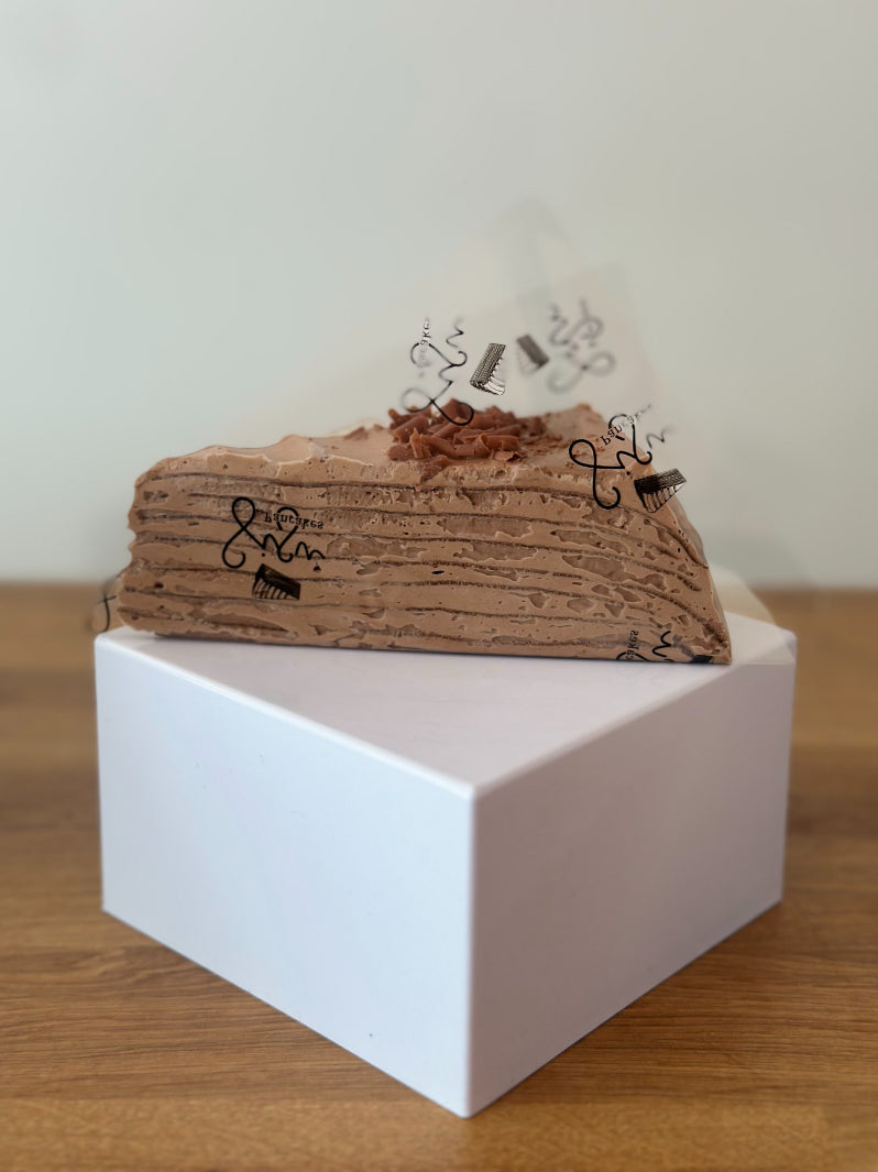 A slice of chocolate pancake ake with chocolate shavings sits on top of a white square box on a wooden table.