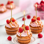 Three cupcakes with pink frosting, raspberries, and lit golden candles on a white surface.