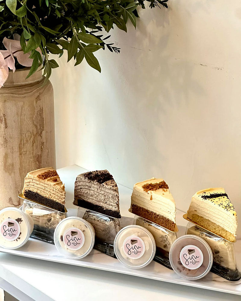 Small slices of a variety of pancake cakes in clear containers on a white surface, with a plant in the background.