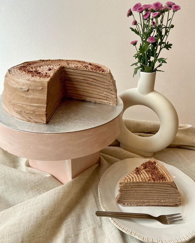 Layered chocolate hazelnut pancake cake on a stand with a slice on a plate, accompanied by a vase of flowers.