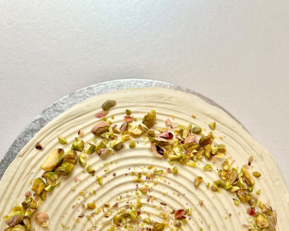 Close up of a pistachio pancake cake with pistachio nuts sprinkled on top of white frosting, set against a white background.