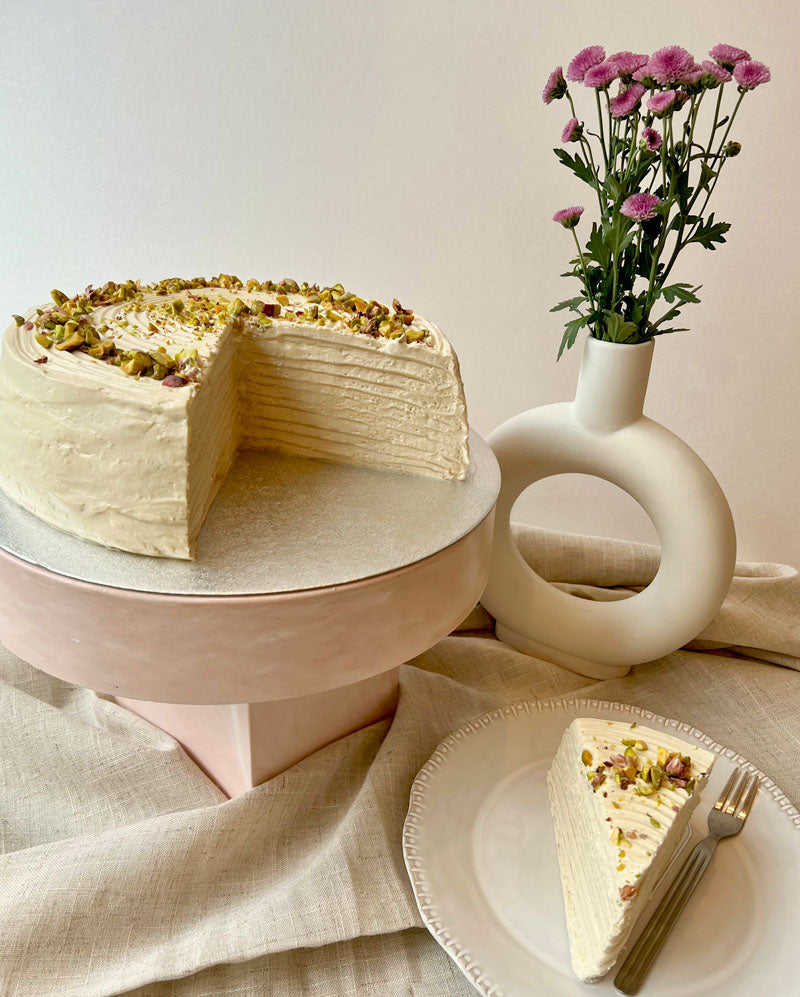 Slice of pistachio pancake cake on a plate with a whole cake and a vase of flowers in the background.