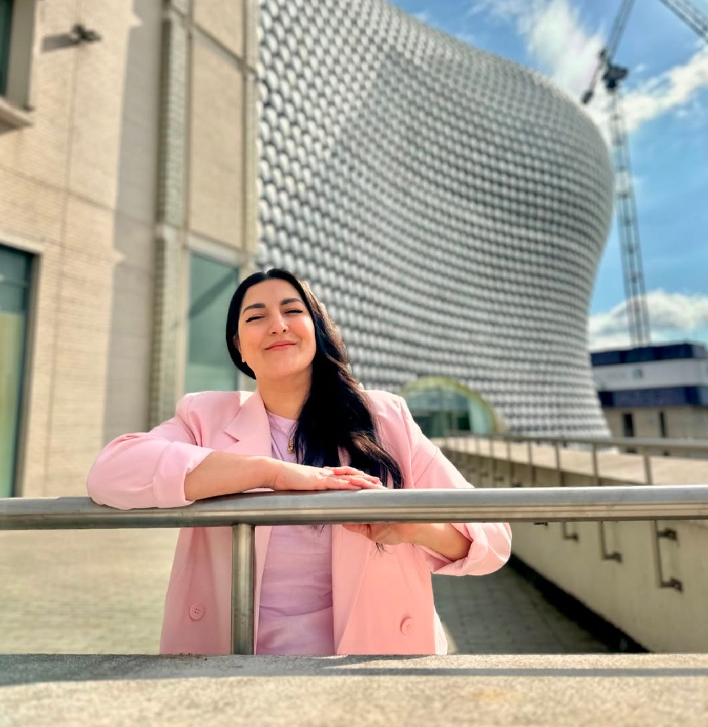 A woman with long dark hair wearing a light pink blazer and matching top leans on a metal railing in front of a modern building with a textured curved facade.