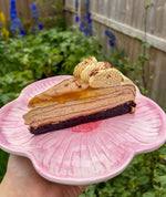 A slice of layered pancake cake on a pink flower-shaped plate, held by a hand in front of a blurred garden background.