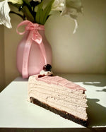 A slice of layered pancake cake with pink frosting and a dark chocolate base, topped with a  cherry, set on a white surface with a pink vase of flowers in the background.