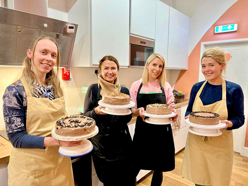 Four women in a kitchen holding cakes on cake stands.