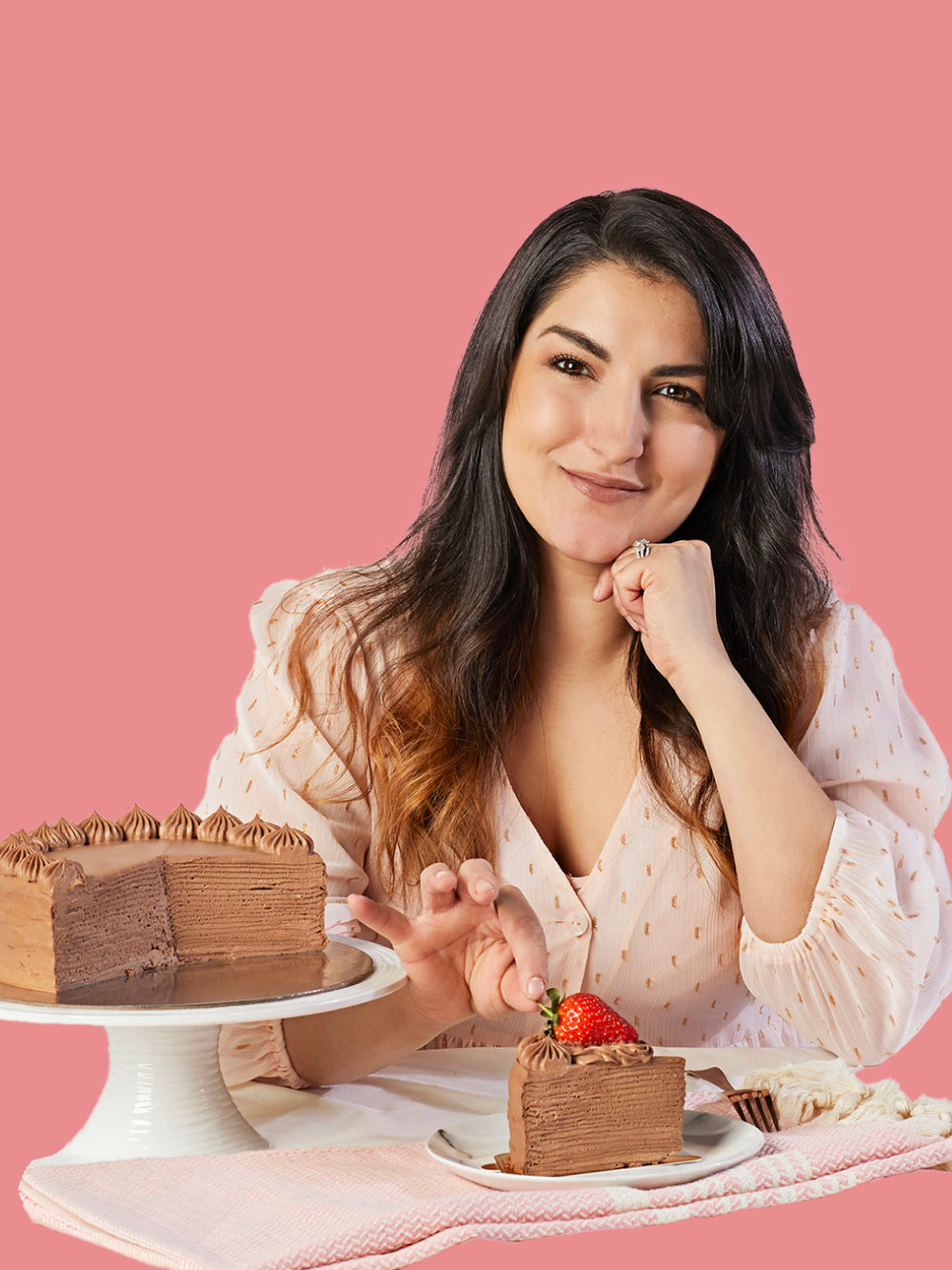 A woman with long dark hair sits behind a table with a chocolate pancake cake on a white pedestal and a slice of chocolate cake in front of her, set against a solid pink background.