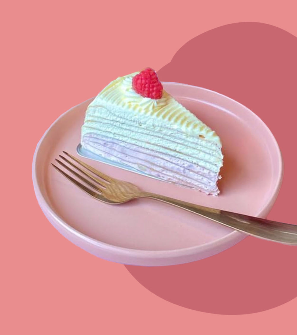 A slice of layered pancake cake with white and pale green frosting on a pink plate, topped with a raspberry, with a gold fork on a pink background.