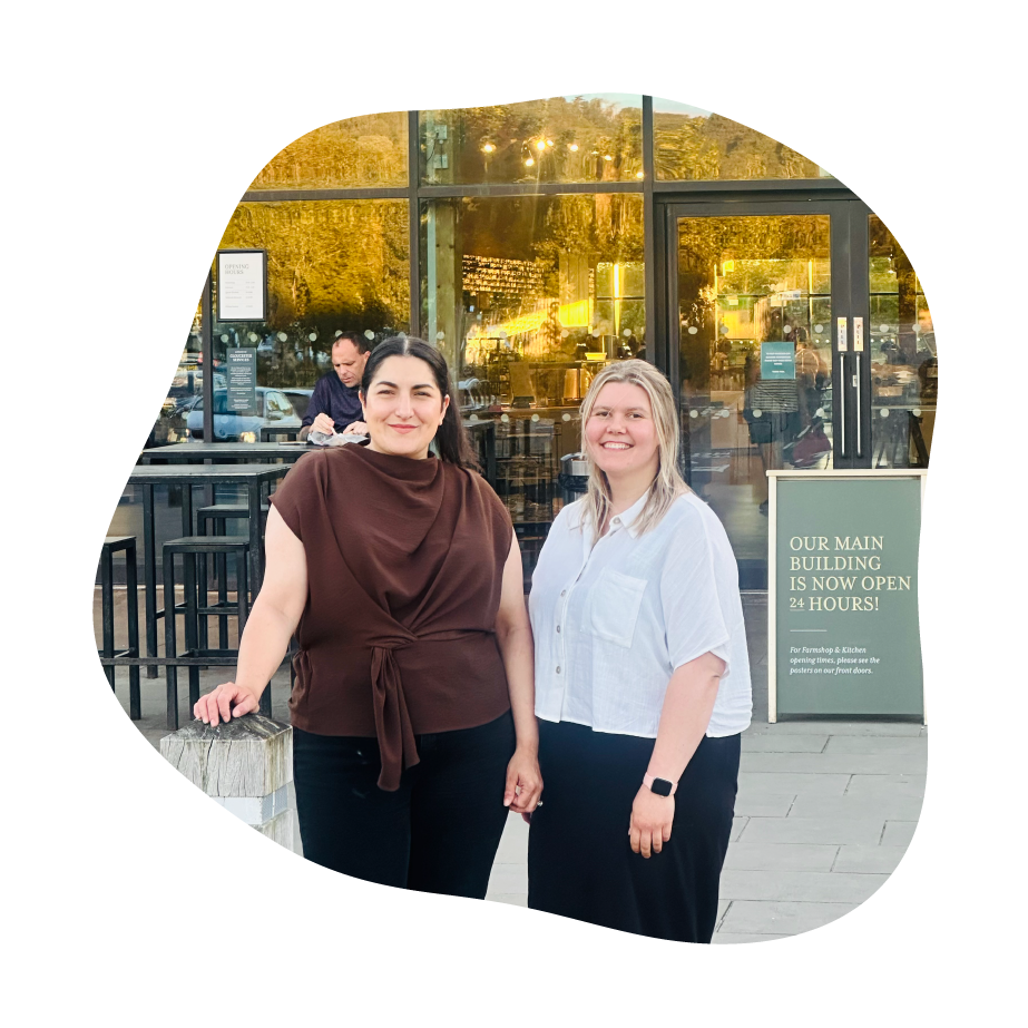 Two women standing in front of a storefront with large glass windows and warm interior lighting. The woman on the left wears a brown draped top and black pants, and the woman on the right wears a light button-up shirt and dark pants.