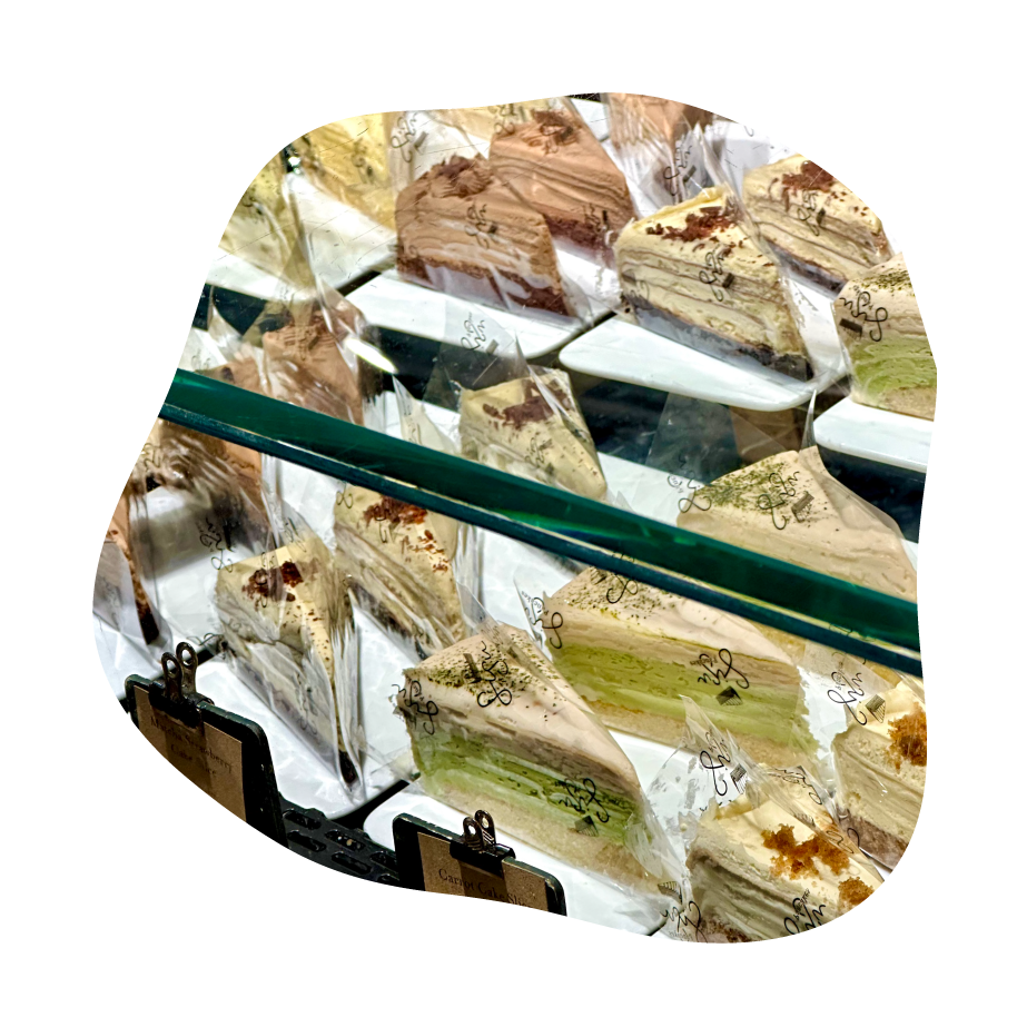A close-up view of multiple pancake cake slices displayed in a glass bakery case, with some wrapped individually in clear plastic. The cakes show cream-colored frosting and layers of green, tan, and brown.