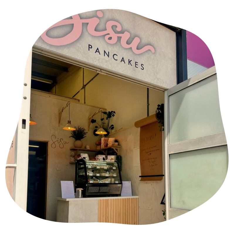 A storefront view of a pancake shop with a large sign reading 'Sisu Pancakes' in pink and black. Inside, a display case with pancakes, shelves with plants, pendant lights, and a wooden counter are visible.