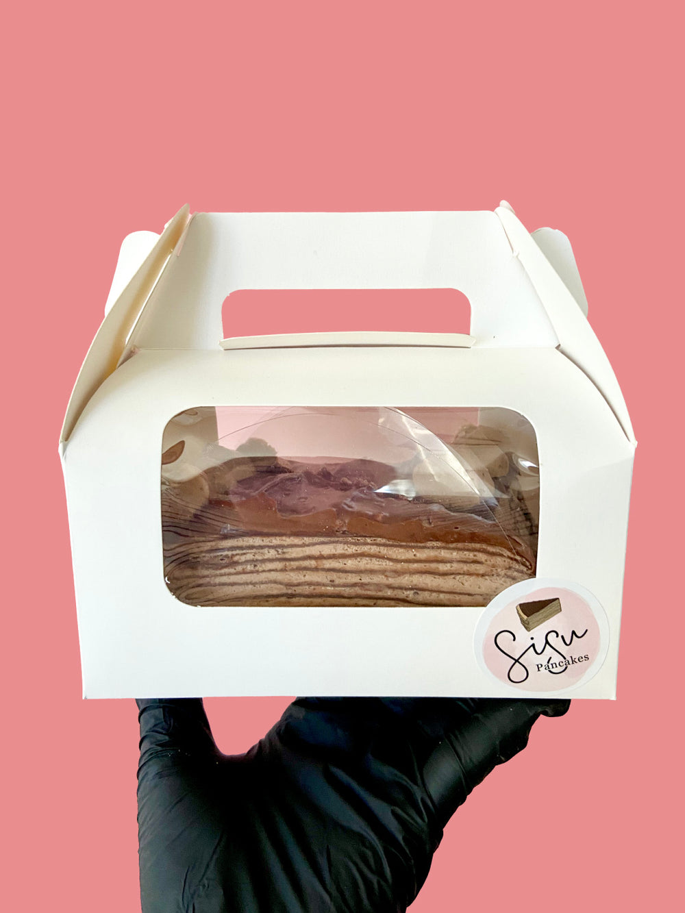 A hand wearing a black glove holds a white cardboard cake box with a clear window showing pancake cake slices inside, against a solid pink background.