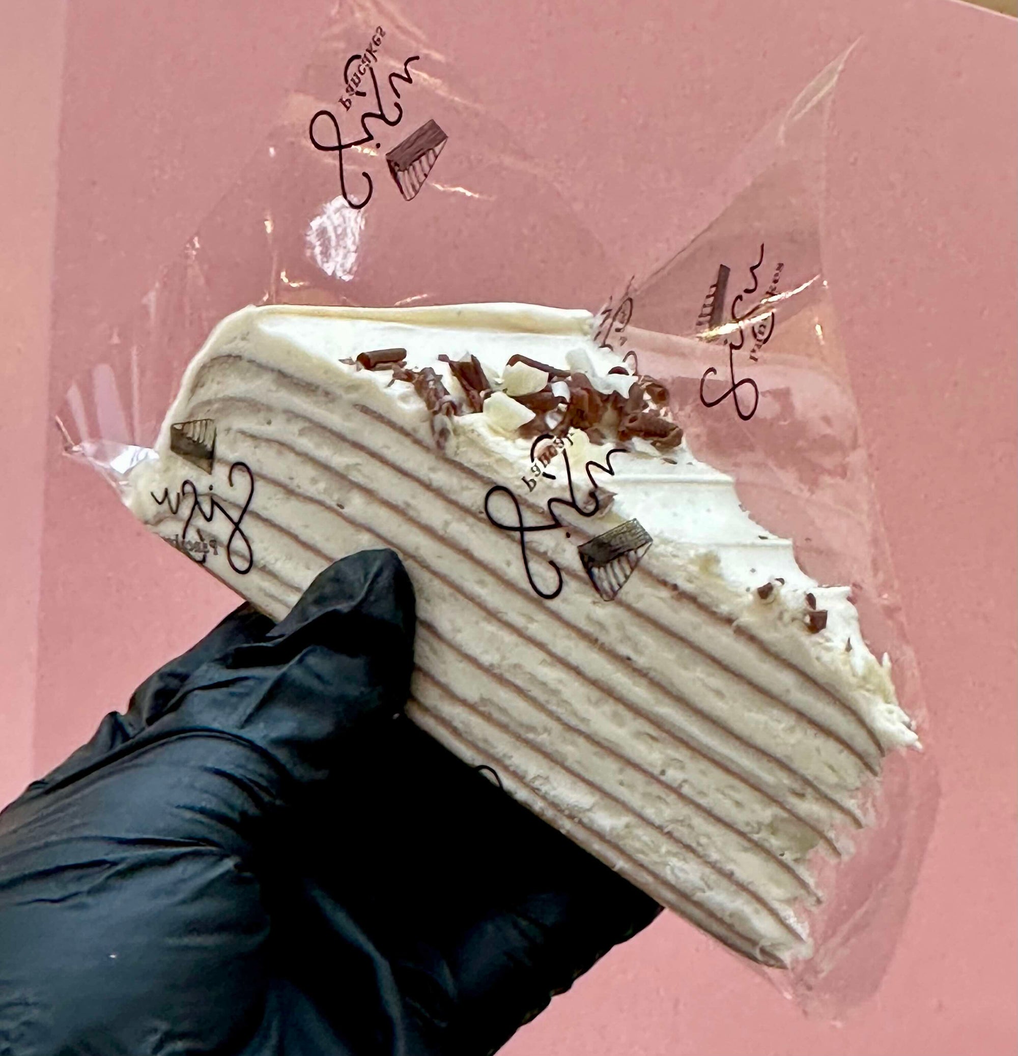 A hand wearing a black glove holds a slice of layered pancake cake wrapped in clear plastic packaging, set against a solid pink background.