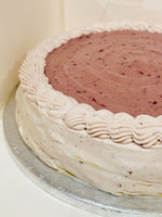 A round pancake cake with a pink cherry top and piped white frosting border, sitting on a silver cake board.