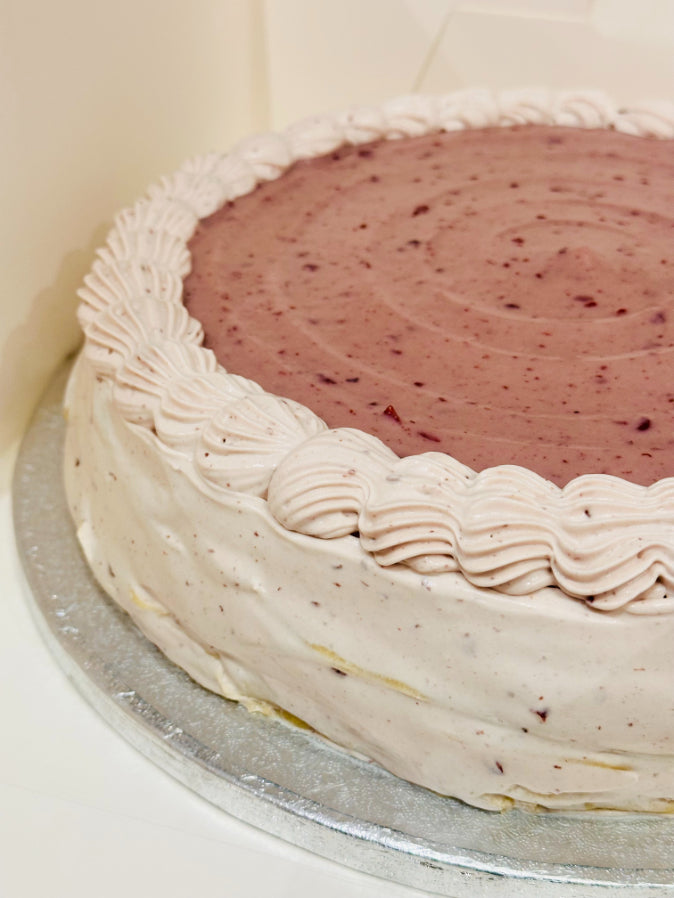 A round pancake cake with a pink cherry top and piped white frosting border, sitting on a silver cake board.