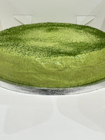 A round green matcha pancake cake with a fine dusting of matcha powder on top, presented on a silver cake board against a white background.