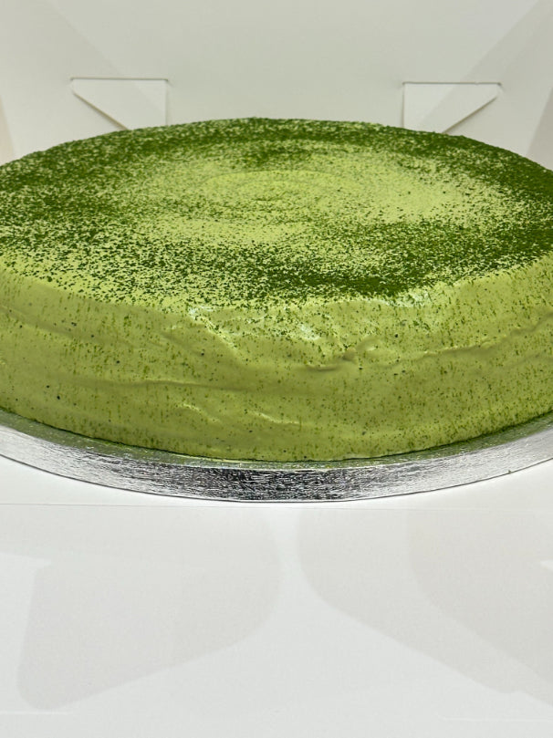 A round green matcha pancake cake with a fine dusting of matcha powder on top, presented on a silver cake board against a white background.