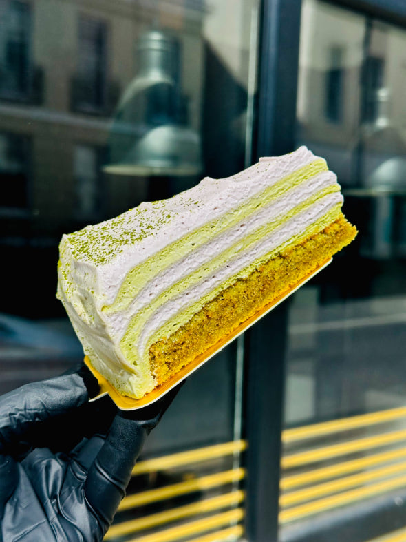 A slice of pancake cake with white and green frosting, topped with green powder, held by a hand wearing a black glove in front of a blurred storefront window.