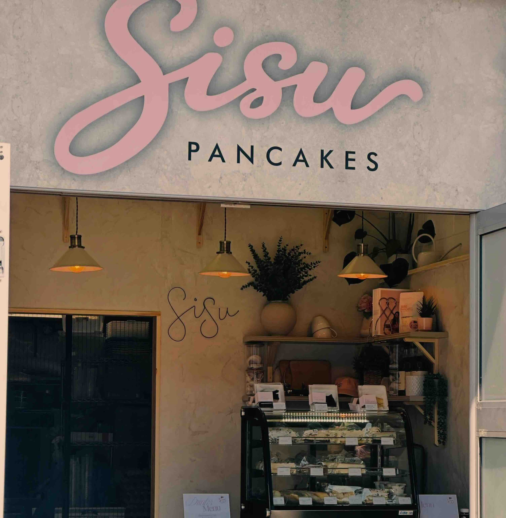 Sisu Pancakes storefront with a large pink script sign reading 'Sisu' and smaller black text 'PANCAKES' above a display case inside a warm beige bakery interior.