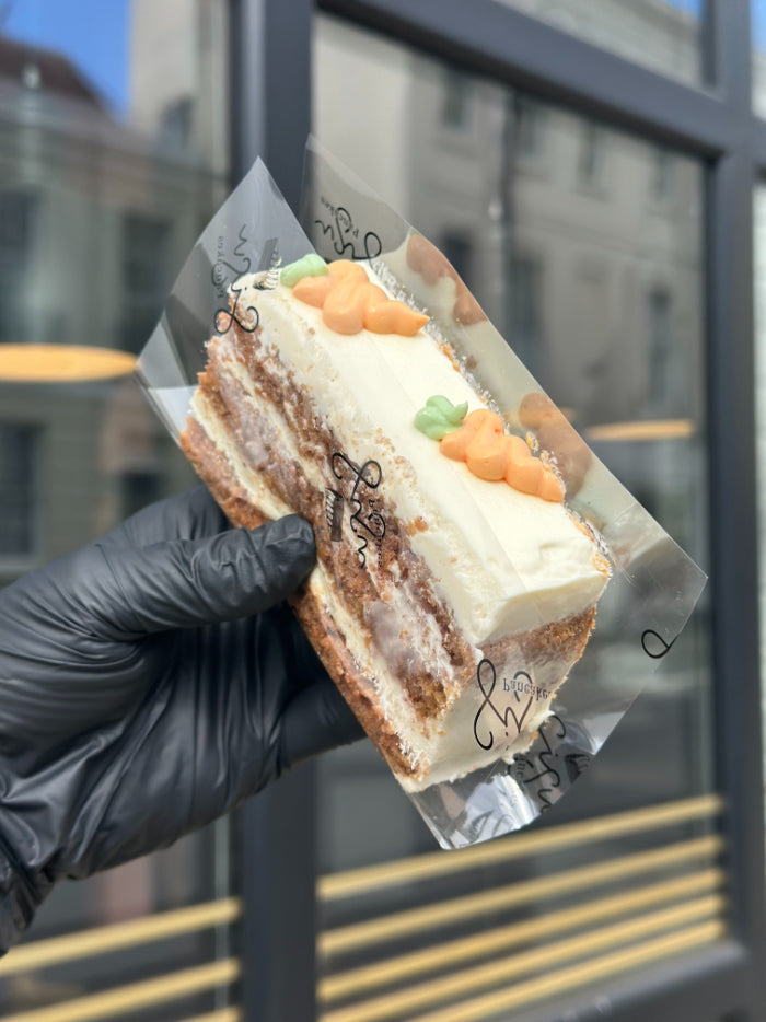 A hand wearing a black glove holds a slice of layered carrot cake with white frosting and small carrot decorations, wrapped in clear plastic in front of a window.