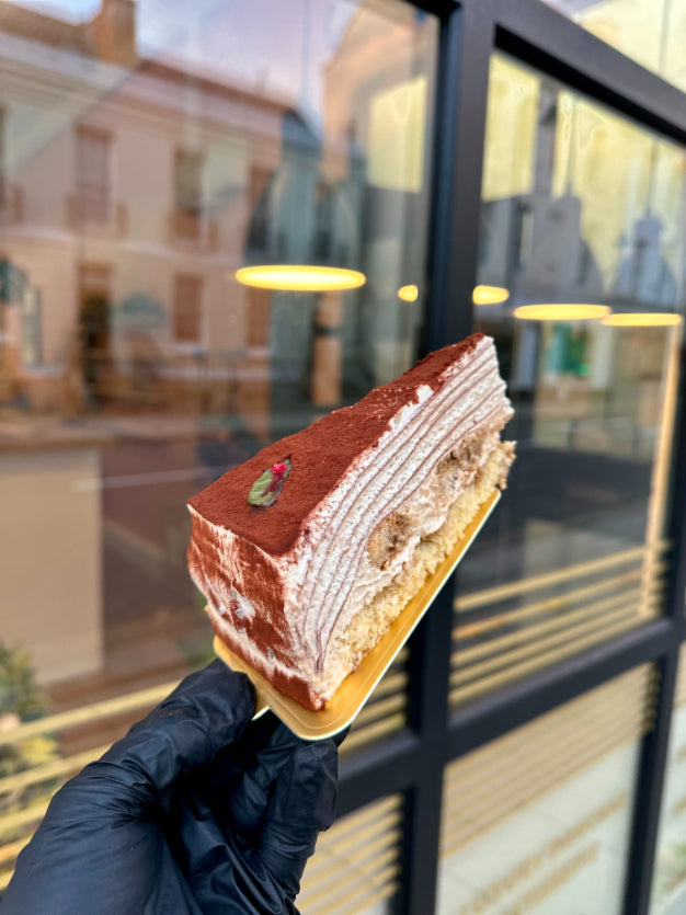 A slice of tiramisu pancake cake, held by a hand wearing a black glove in front of a glass storefront window with reflections.